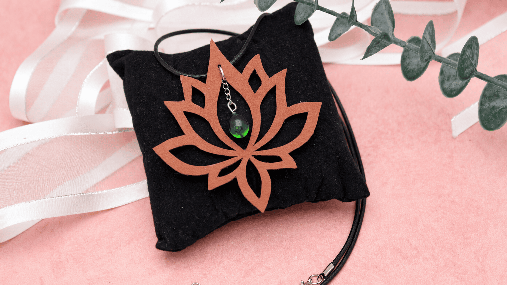 Collier artisanal lotus en cuir recyclé avec petite goutte verte en verre recyclé, posé sur coussin noir sur fond rose poudré.